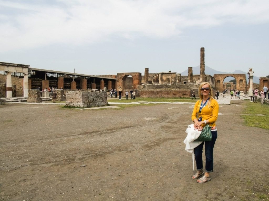 Lindsey poses in front of the ruins of Pompeii, wearing jeans and a white cardigan, carrying a bag and jacket.