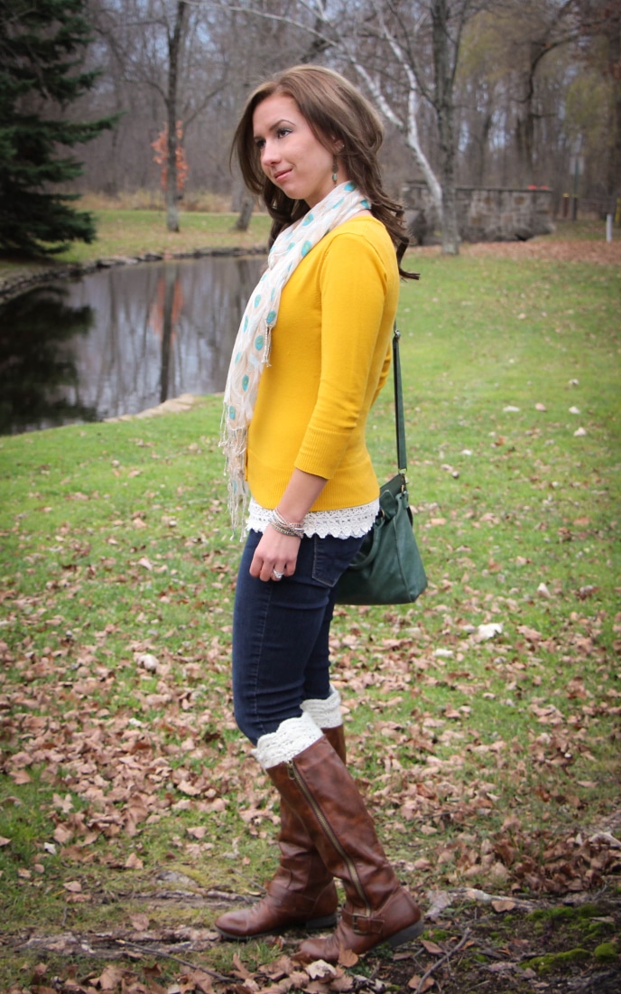 Lindsey poses in front of a pond at a park on a sunny Fall day. She's wearing jeans paired with a yellow cardigan and white scarf, knee-high boots, and a cross-body bag.