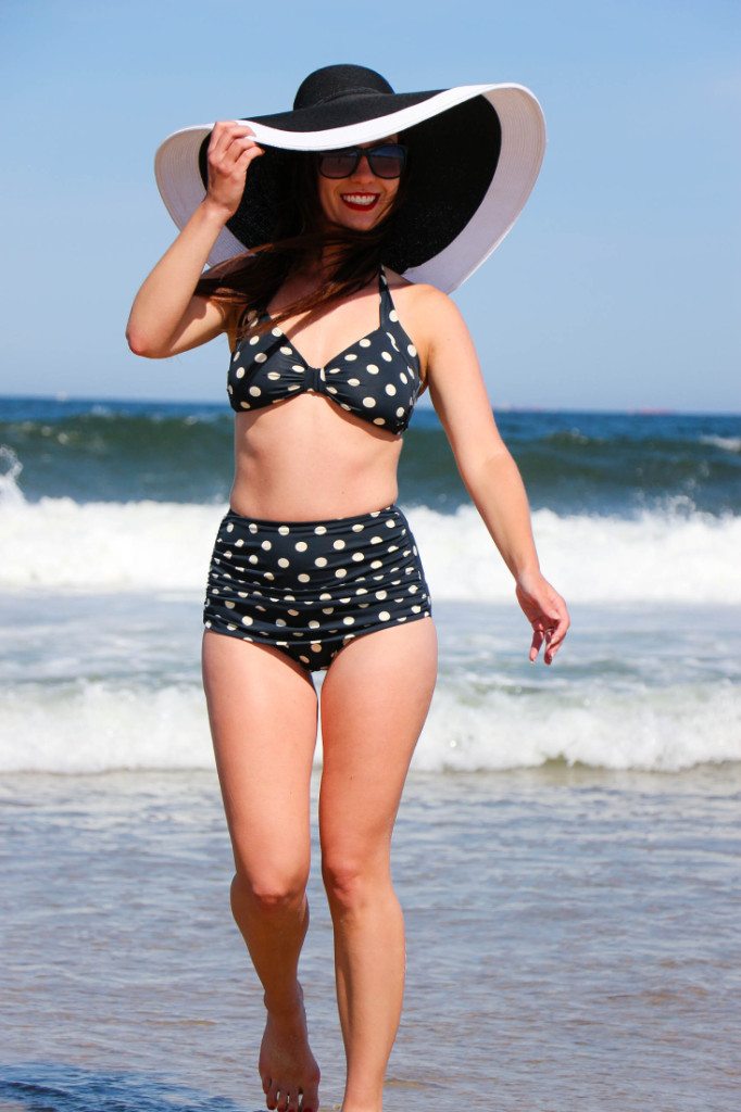 A woman wears a black and white polka dot bikini and a black and white floppy sunhat. She walks along the waves of a beach in the retro-style high-waisted bikini with her hand on her hat. 