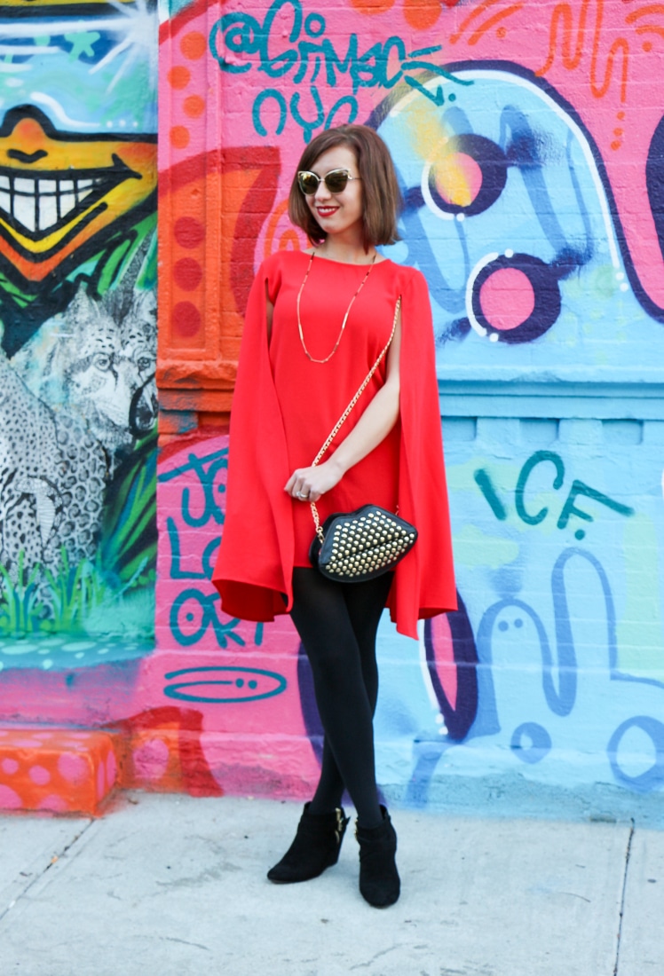 A woman with short dark hair standing against a bright graffiti wall in New York City wearing a Shein Red Cape, black tights, and black ankle boots