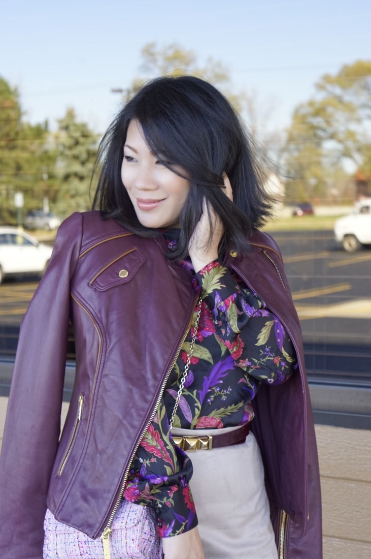 A woman poses outdoors in a stylish fall outfit. She wears a colorful patterned sill shirt under a deep maroon leather jacket, paired with khaki pants.