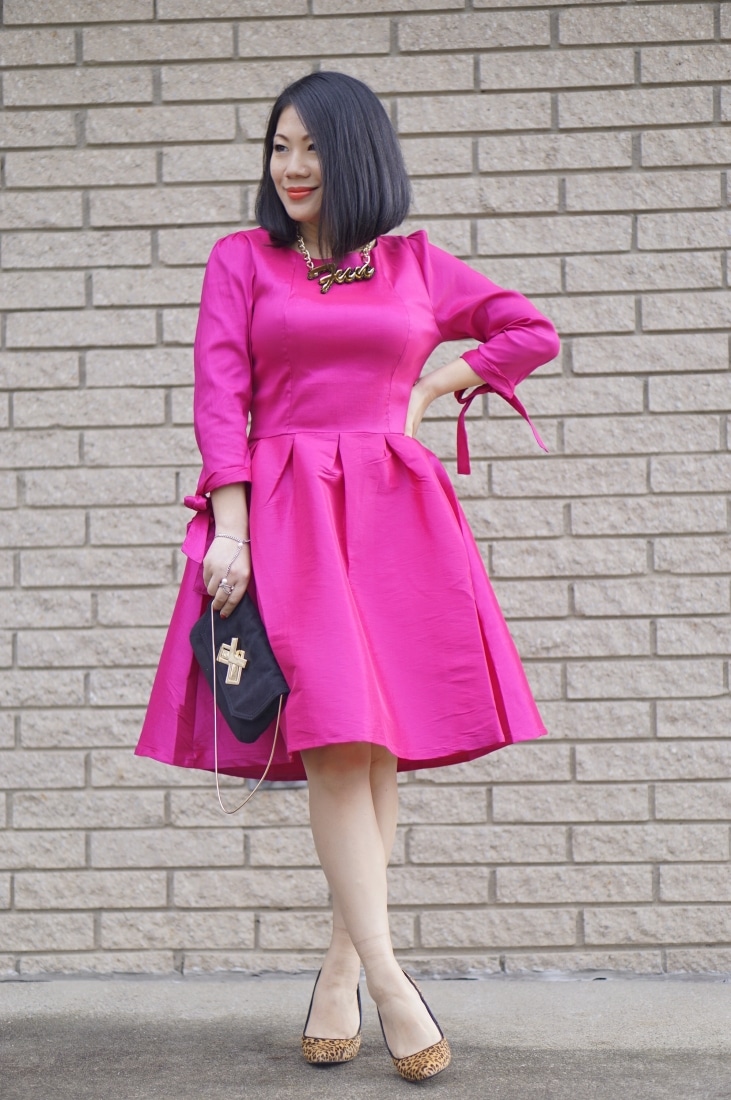 A woman poses in front of a tan brick wall. She's wearing a vibrant long-sleeved link dress, holding a black handbag, and wearing cheetah print high-heeled shoes.