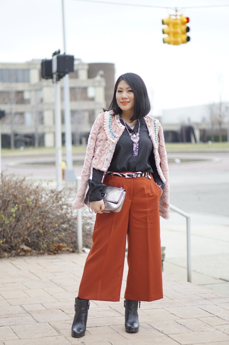 A woman poses outside, showing off her fall-inspired outfit. She wears burnt-orange cropped pants with black heeled boots, a black silk blouse under a couture pink jacket.