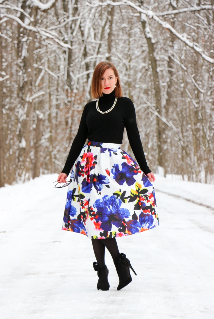 Lindsey wearing a black turtle with colorful floral midi skirt, black tights, and black bow heels standing on snow covered path in the woods