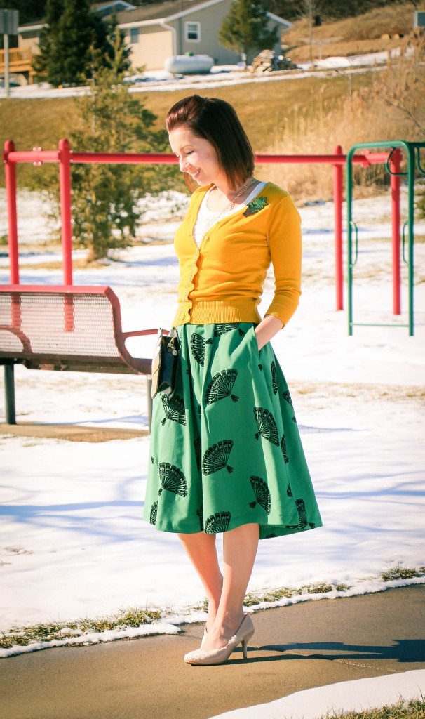 Lindsey poses in front of a snowy playground. She's wearing a yellow buttoned up cardigan and green skirt with an all-over fan pattern, paired with nude heels.