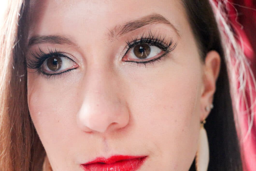 A closeup image of a woman's face. She's wearing makeup -- red lipstick, foundation, shimmery eye shadow, and eyeliner. She has full, wispy eyelash extensions.