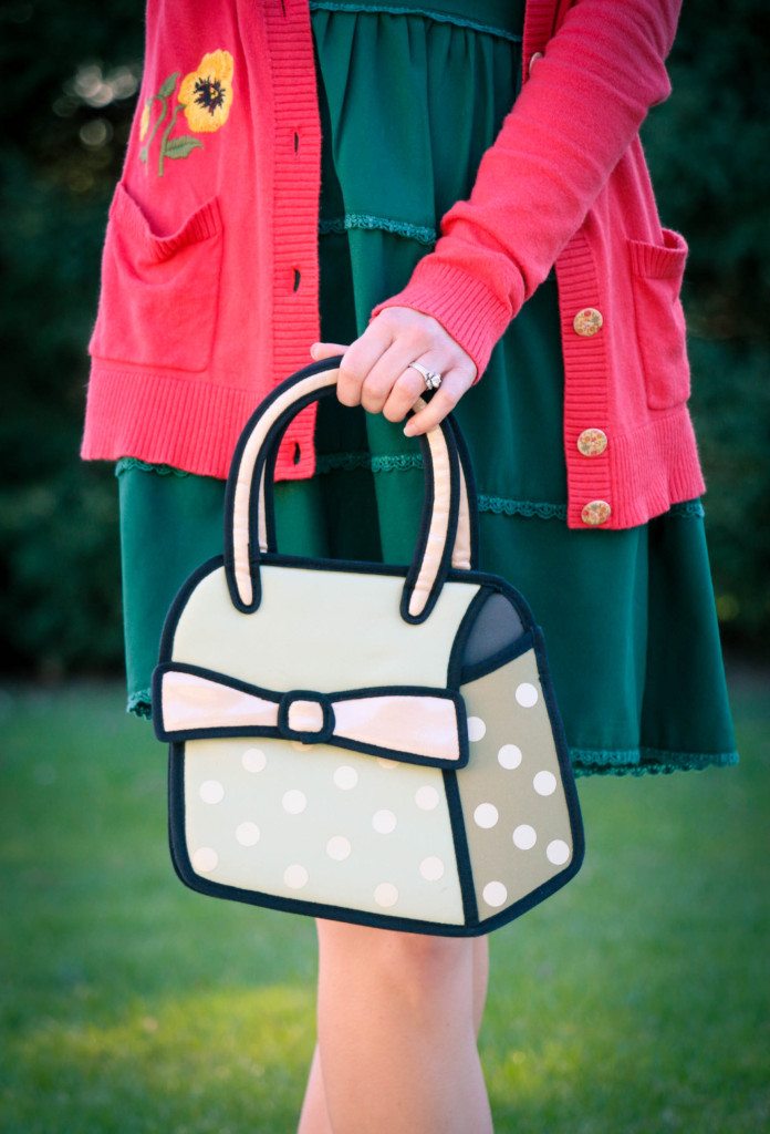 A woman holds a novelty handbag that's designed to look like a 2D comic illustration. The purse is a light mint color with white polka dots and a light pink bow.
