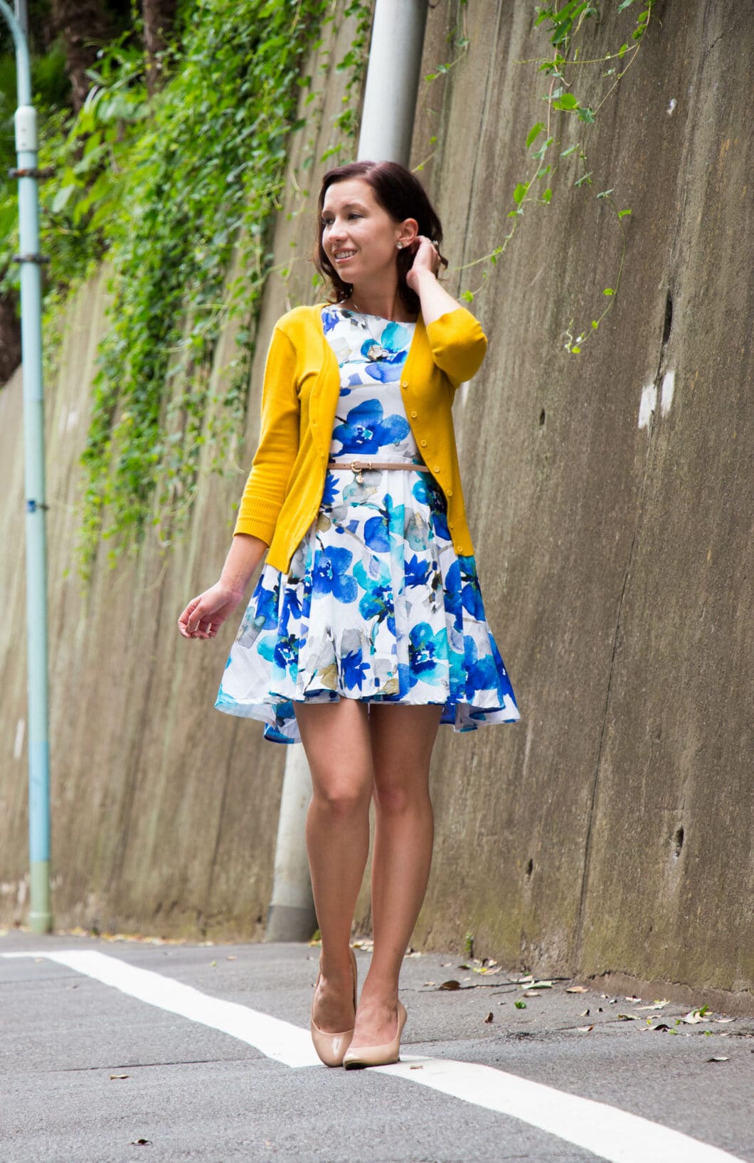 Lindsey poses on a city street wearing a blue and white flower patterned dress paired with a yellow cardigan.