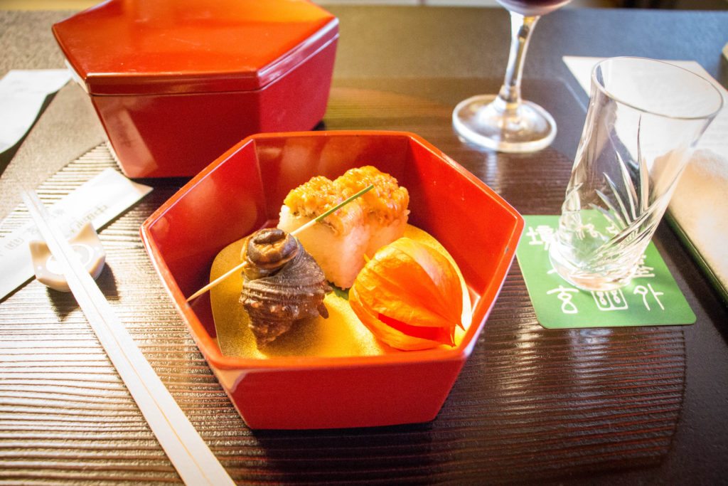 A hexagon-shaped red bowl holds multiple pieces of sushi, and chopsticks rest on the table next to the bowl