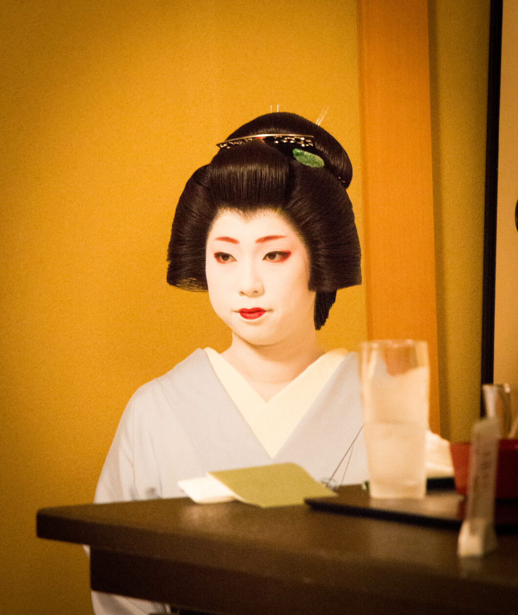 A young Japanese woman dressed as a traditional Geisha kneels in front of a yellow wall.