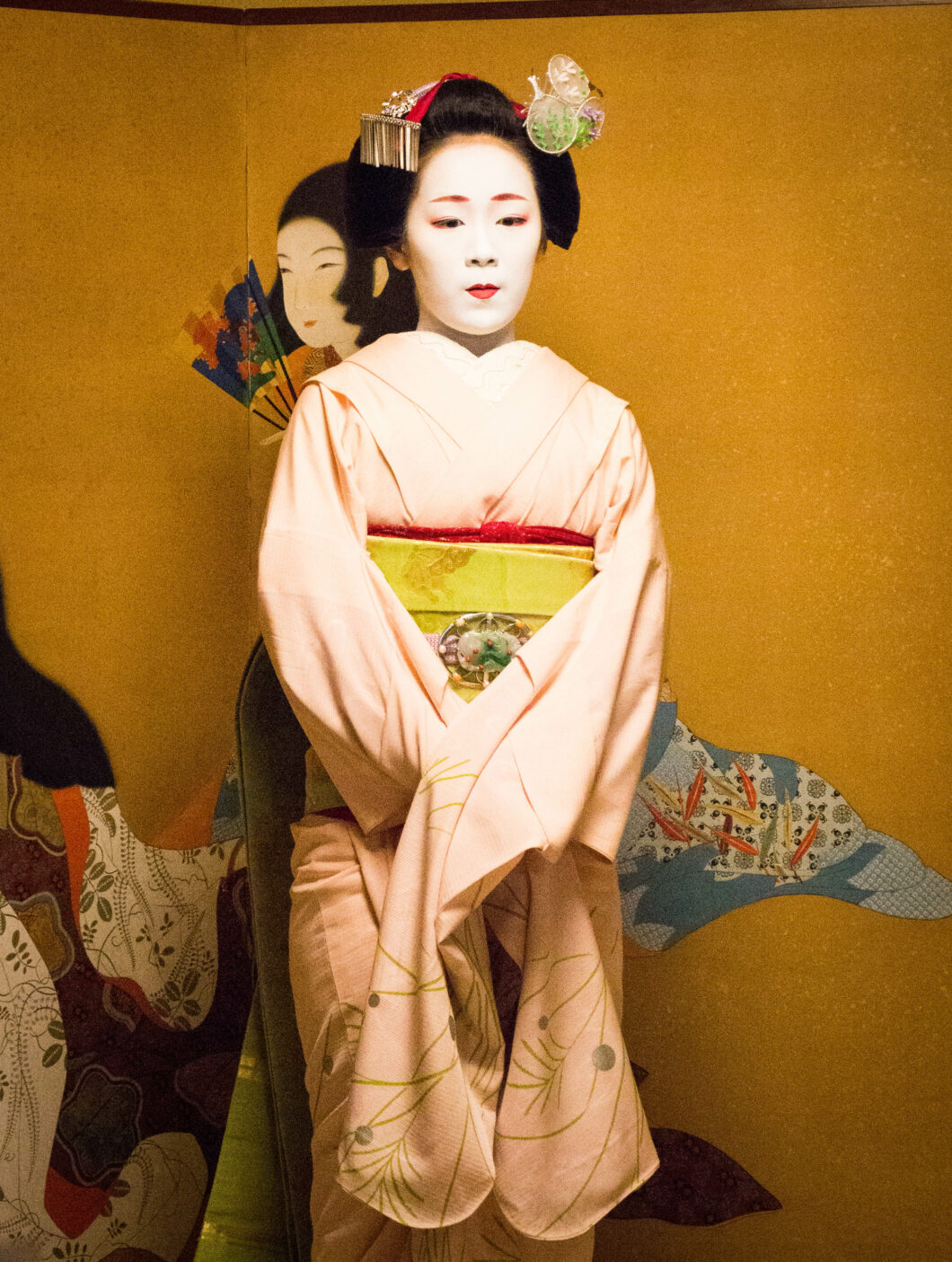 A young, stoic-faced Japanese Maiko stands against a yellow background. She's wearing traditional Geisha makeup and clothing.