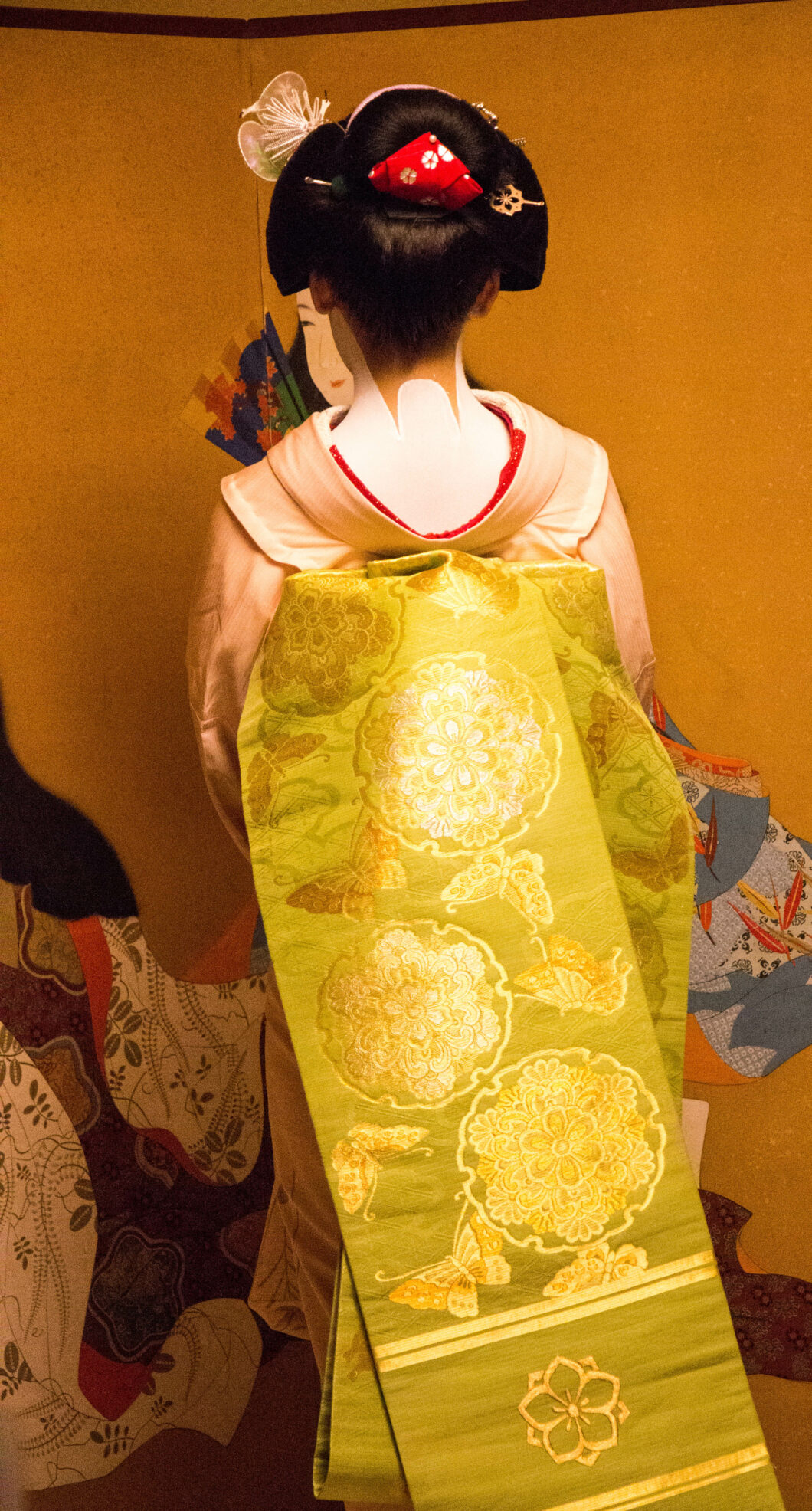 A Japanese Maiko stands with her back to the camera, showing the traditional sash garment called and Obi. The garment is a stunning gold color with intricate patterns.