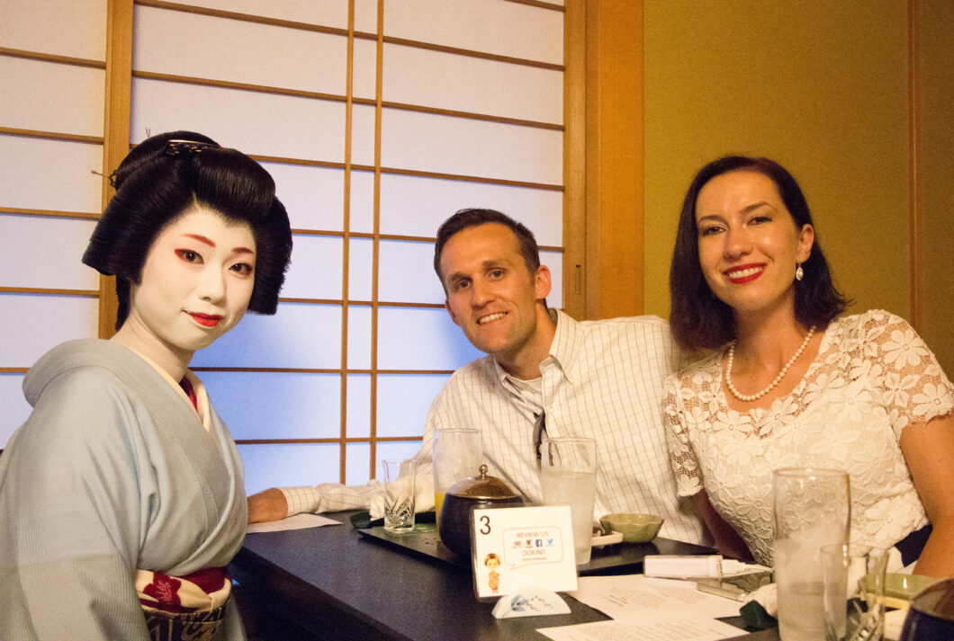 An American man and woman sit at a small table across from a Japanese Geisha. They're enjoying tea together.