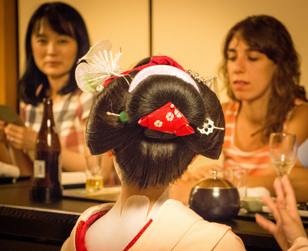 The back of a young Geisha's head shows a traditional Geisha hairstyle -- the woman's long brown hair is tightly pulled back in a neat bun that resembles a bow, and is accessorized with hair red and white hair clips.