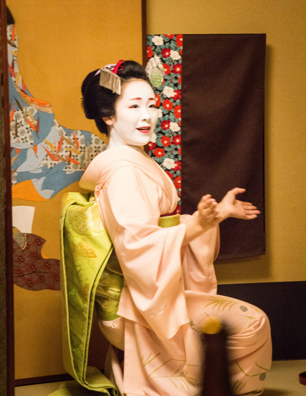 A young Maiko kneels on one knee, clapping along with music during a dinner performance. She is smiling and looking at the guests.