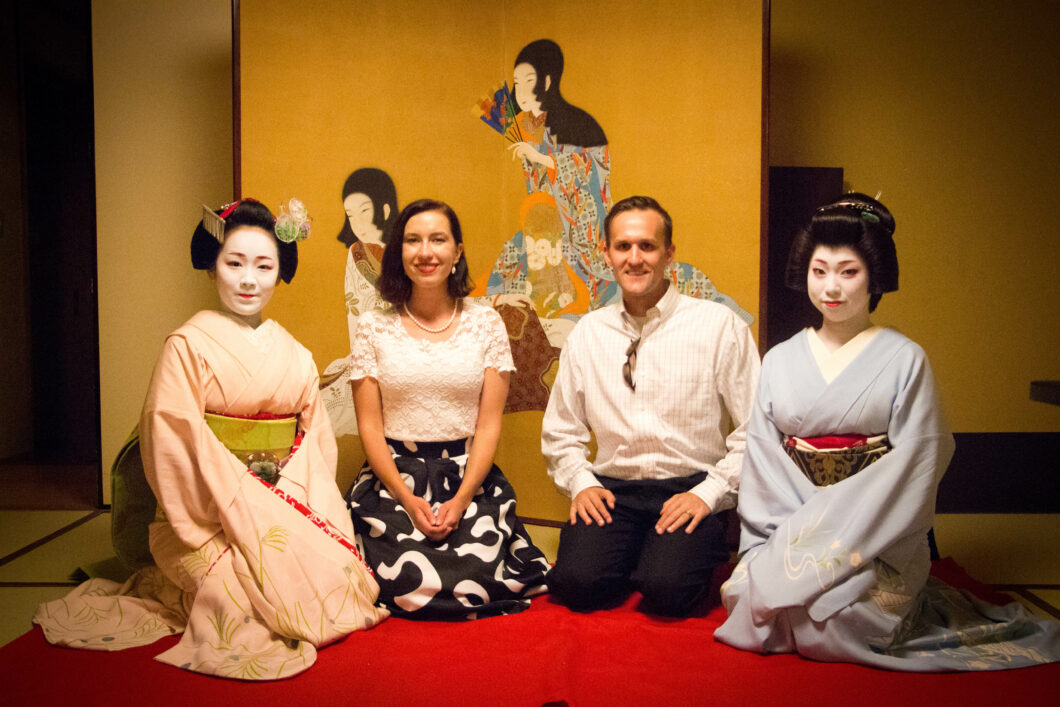 An American man and woman pose with two young Japanese geishas. The man and woman are in the middle, with a Geisha on either side of them. Everyone is kneeling on a red carpet in front of a screen with Japanese artwork.