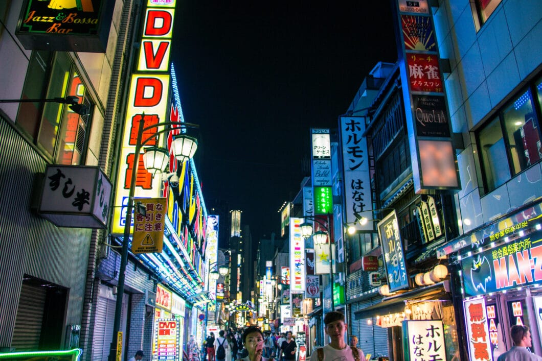 A busy city street in Japan at night, lined with brightly-lit storefront signs advertising shops and DVD stores.