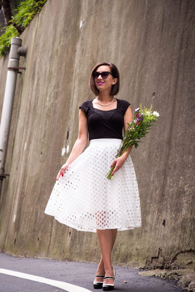 A woman holding a fresh flower bouquet poses in a black and white outfit; she's wearing a knee-length white midi skirt, black cap-sleeve top, sunglasses, and heels.