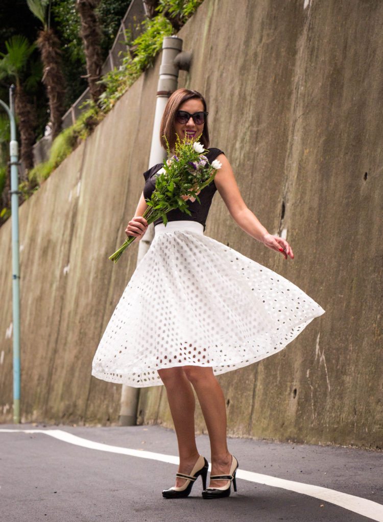 A woman twirls as she stands on a street. She's wearing a black short-sleeve top, white eyelet midi skirt, and black and white Mary Jane heels. She's holding a fresh flower bouquet.