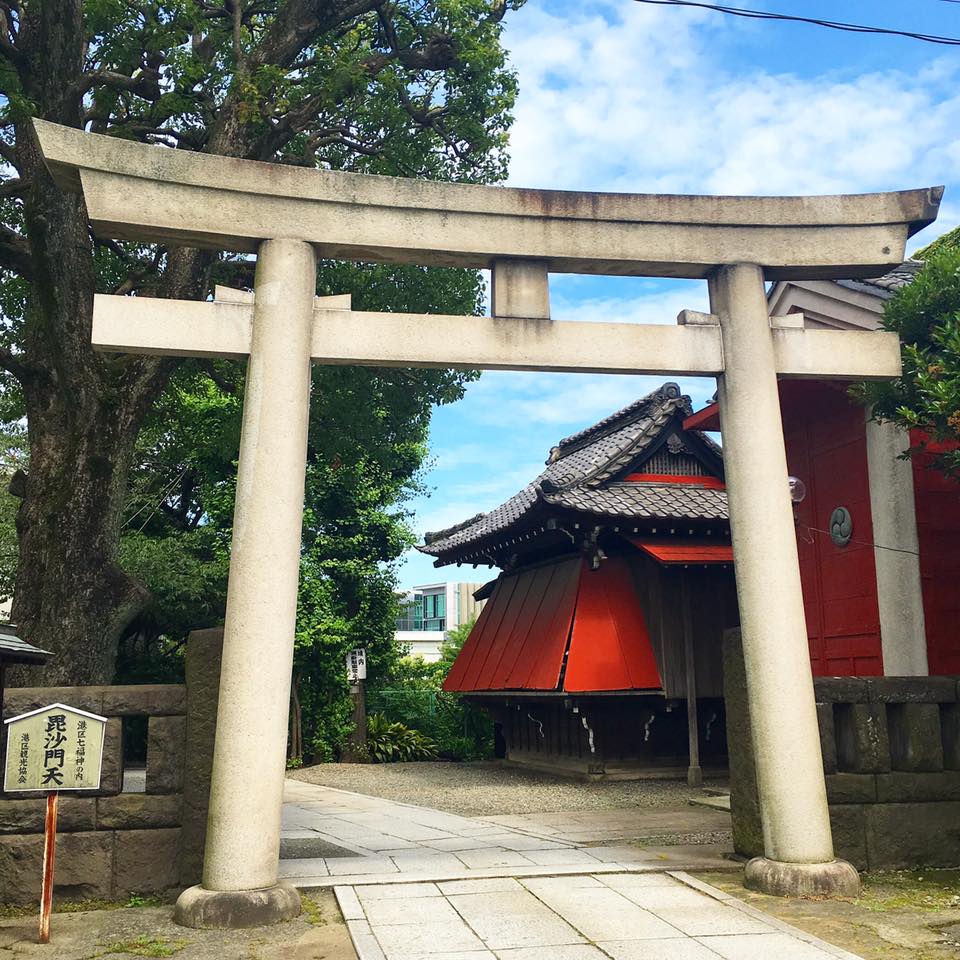 An image shows a large stone shrine in front of an ancient Japanese temple. The temple is bright red and surrounded by green trees under a blue sky.