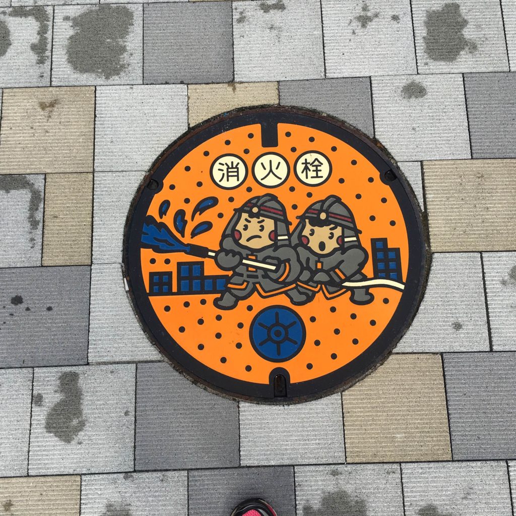 A Kawaii style manhole over in the streets of Japan features a cartoon of two firemen putting out a fire.