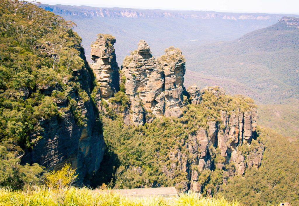 The Three Sisters rock formation in the Blue Mountains of Sydney, Australia, on a clear and sunny day. The rock formation sits on a cliff in a lush green forest valley.