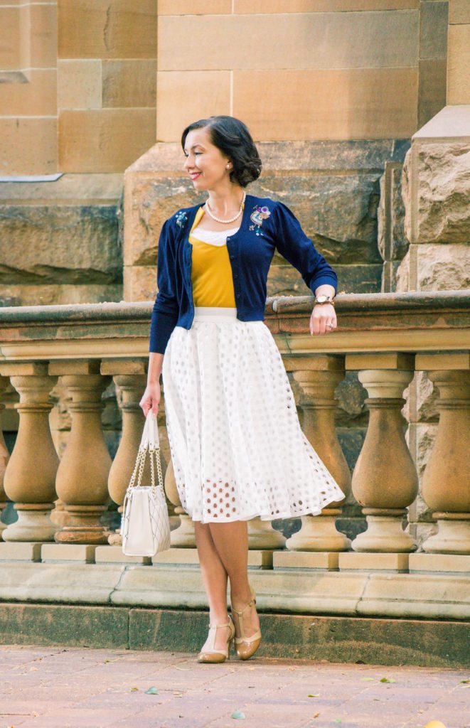 A woman poses against a stone railing, wearing a knee-length white skirt, navy blue sweater, golden-yellow top, and nude t-strap heels.