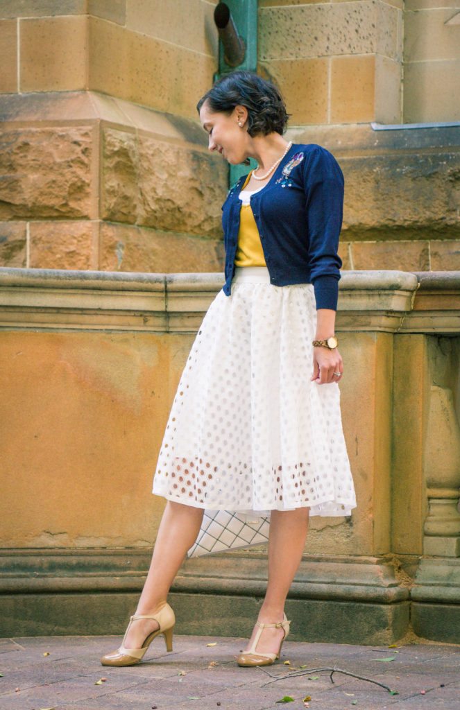 A woman poses wearing a white skirt, navy blue cardigan over a yellow top, and nude t-strap heels.