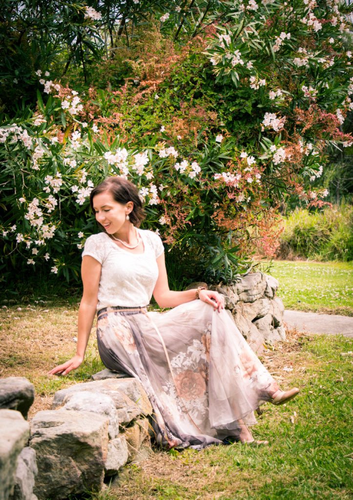 A woman sits on a stone wall in a lush garden. She's wearing a long, flowy floral patterned skirt, white lace top, and pearl accessories.