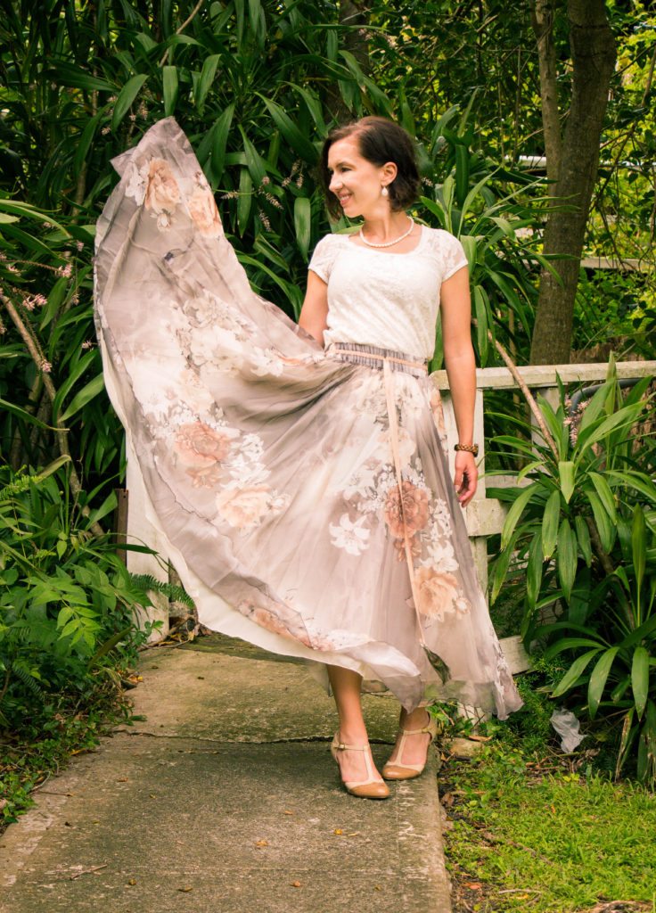 A woman poses in a lush garden scene. She's poses, swishing her long, neutral colored floral skirt in the air. She paired the skirt with nude t-strap heels and a white cap-sleeved shirt.