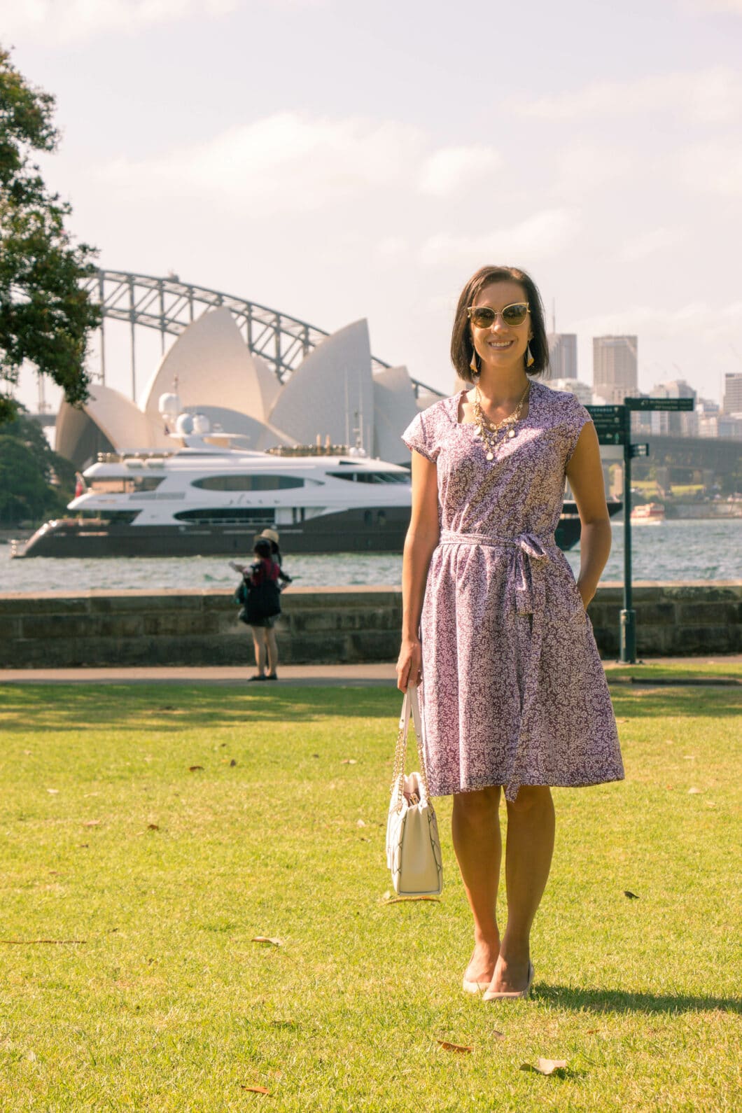 A woman poses on a grassy lawn wearing a summery floral dress and sunglasses. The Sydney Opera House is pictured in the background.