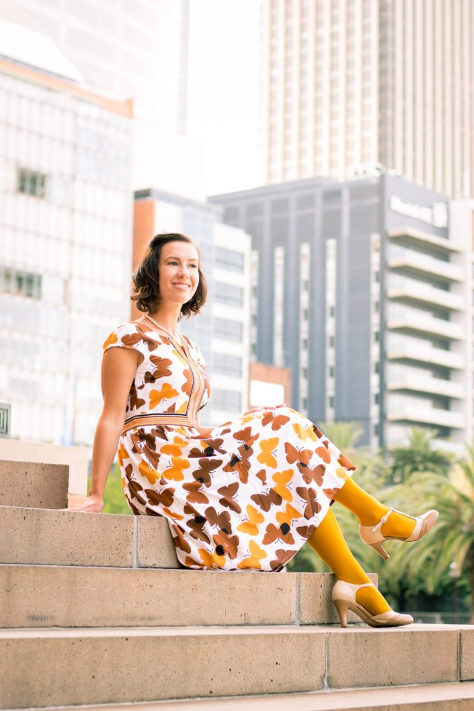 A woman sits on stone steps, wearing a brown and yellow patterned butterfly dress, yellow tights, and nude t-strap heels.