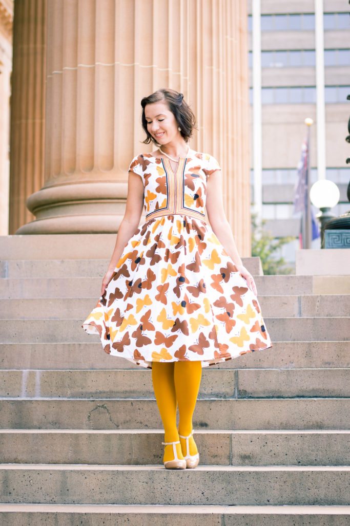 A woman poses standing on stone steps. She's wearing a knee-length dress with an all-over butterfly pattern in shades of brown and yellow, with white tights and nude heels.