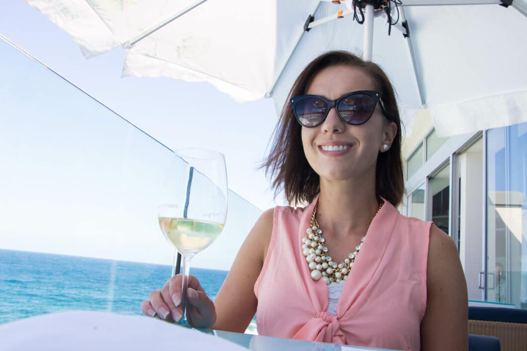 A woman with short brown hair, wearing sunglasses and a pink top, enjoys a glass of white wine at an outdoor restaurant table, overlooking the ocean and clear blue skies.