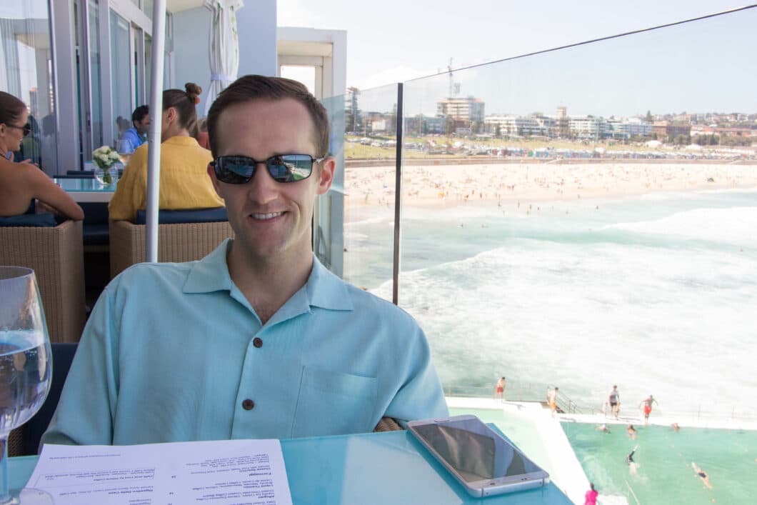 A man wearing a light blue short sleeved collard shirt and sunglasses is seated at a dining table, overlooking Bondi beach at Icebergs in Sydney.