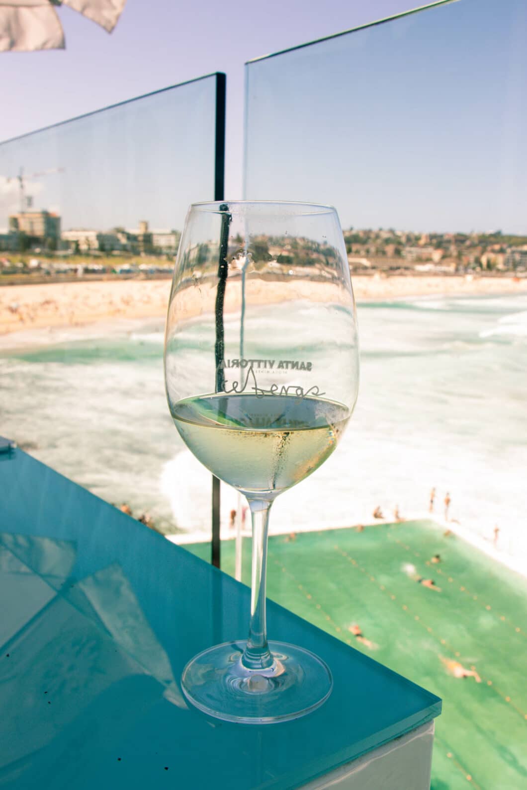 A glass of white win rests on a glass table. In the background, there's a pool that overlooks the ocean and a shoreline filled with people enjoying the clear, sunny day.
