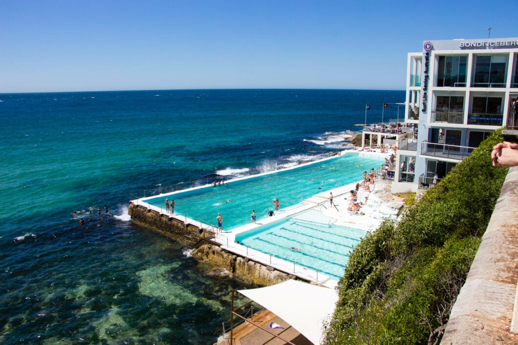 An image overlooking the pool area of the Bondi Icebergs restaurant, which looks out onto the vast ocean. Vacationers swim in the pool and tan on chairs.