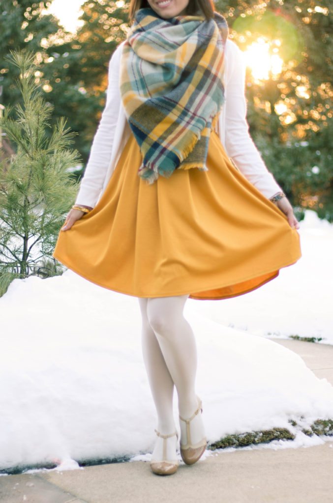 A woman stands on a snowy sidewalk, showing off her wintery yellow long sleeve dress paired with a yellow plaid scarf, white tights, and vintage heels.