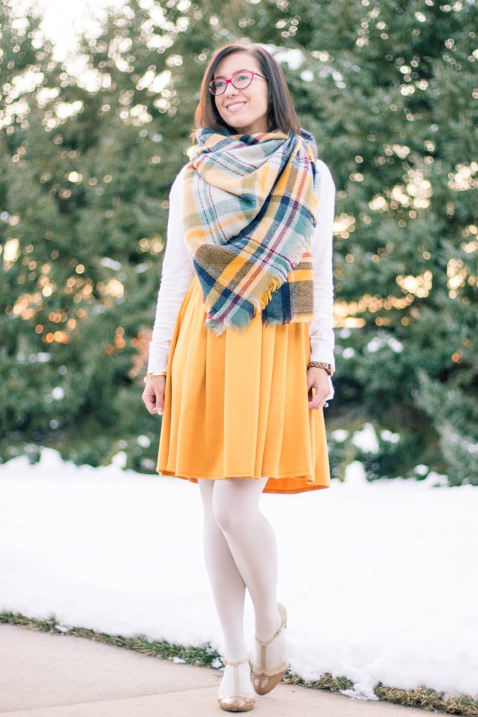 A woman poses on a snowy sidewalk. She's wearing a yellow dress with white long sleeves, a yellow and blue plaid scarf, and white tights, paired with a pair of nude t-strap heels.