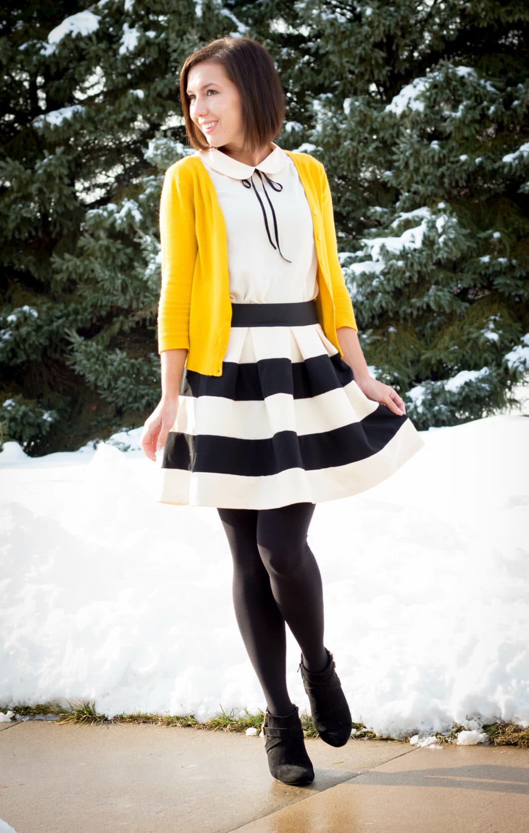 Lindsey wearing a striped mini skirt, cream blouse with a yellow cardigan, black tights, and ankle boots