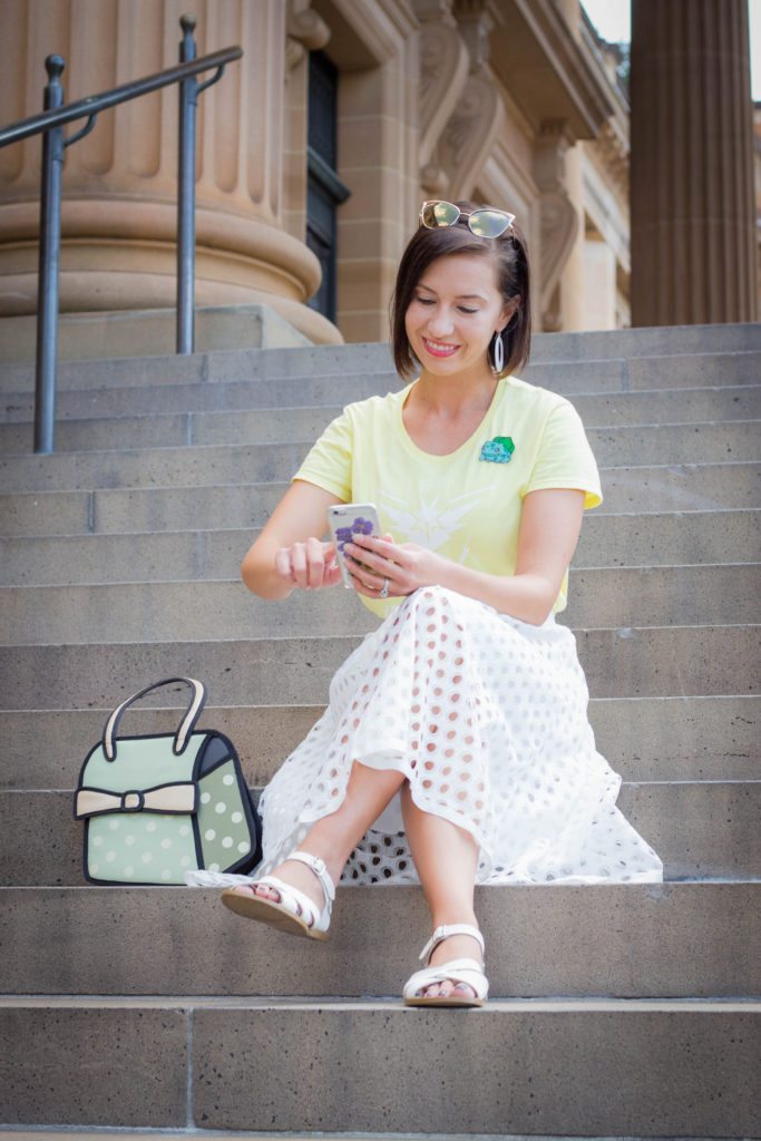 A woman sits on stone stairs, wearing a white eyelet skirt and a yellow shirt, holding her phone with sunglasses on her head and a stylish purse sitting on the stairs next to her.
