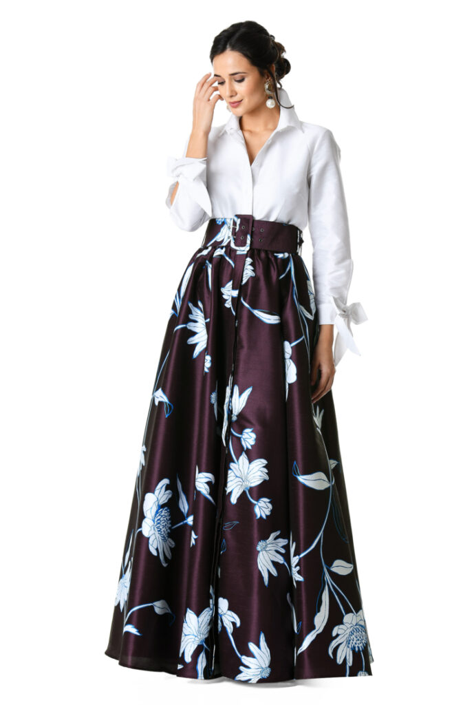 A woman wears a long, floor-length skirt with an elegant flower pattern, paired with a long-sleeve white blouse.