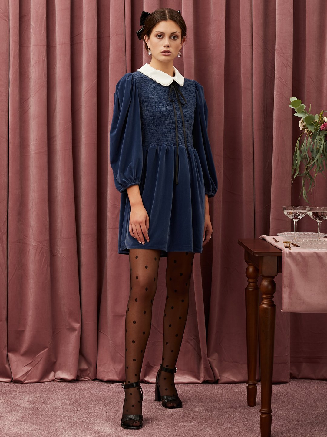 A model poses wearing a cocktail-length navy blue velvet mini dress, with long sleeves and a white collar. She stands in front of a wall of mauve velvet curtains.