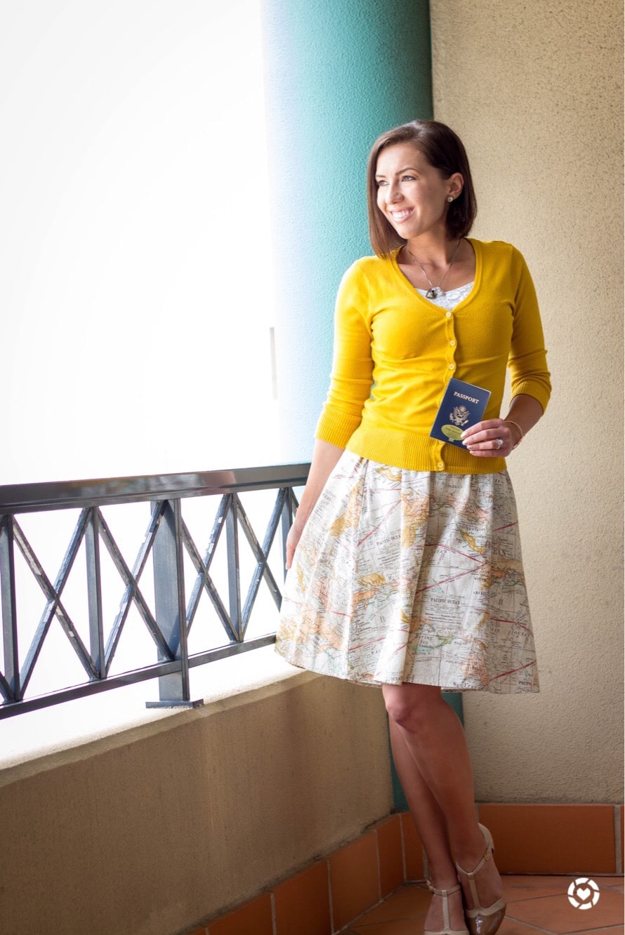 A woman poses on an outdoor balcony. She's wearing a patterned dress with a map of the world on it, and a buttoned-up yellow cardigan sweater. She's holding a US Passport.