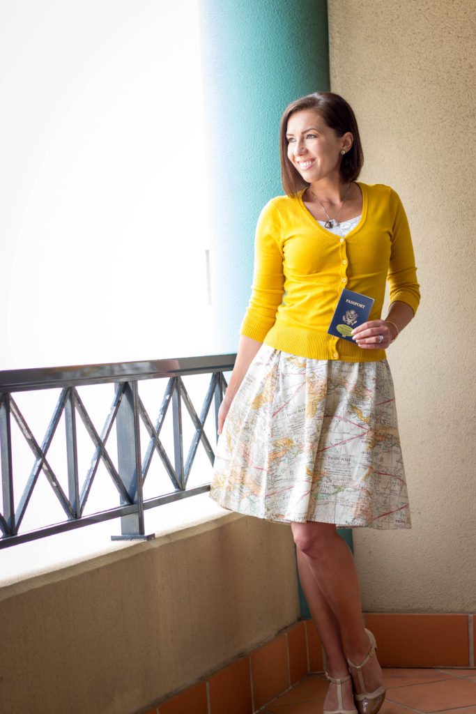 Lindsey poses on a hotel balcony, holding her passport. She's wearing a yellow buttoned-up cardigan and a white skirt with an all-over map pattern.