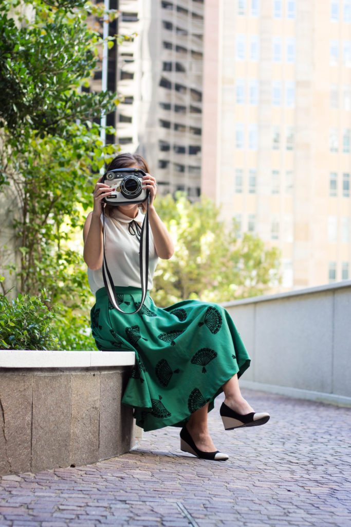 A woman sits on a bench, holding a novelty purse shaped like a camera up to her face, mimicking taking a picture.