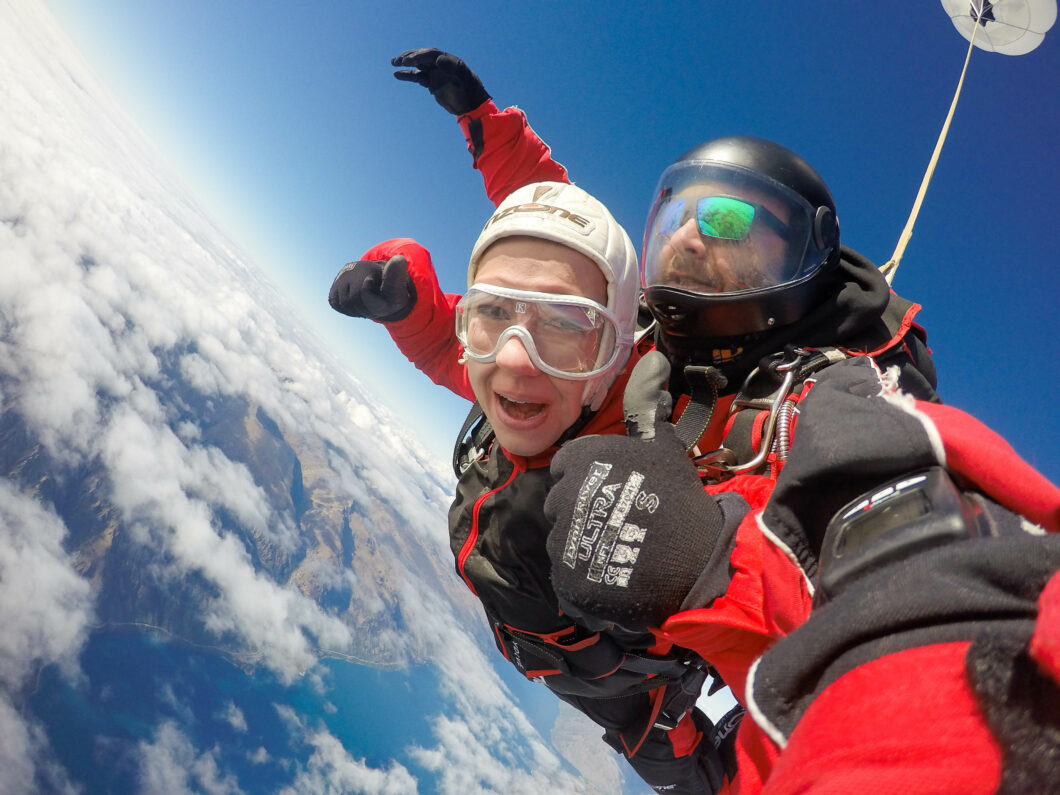 Lindsey tandem skydiving in New Zealand, smiling with thumbs up above the clouds 