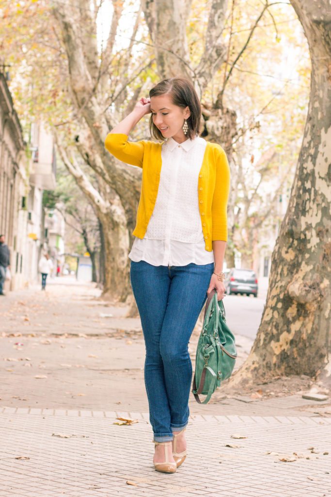 A woman casually poses on a city sidewalk. She's wearing blue jeans, a white top, and yellow sweater, paired with a green handbag and nude t-strap heels.