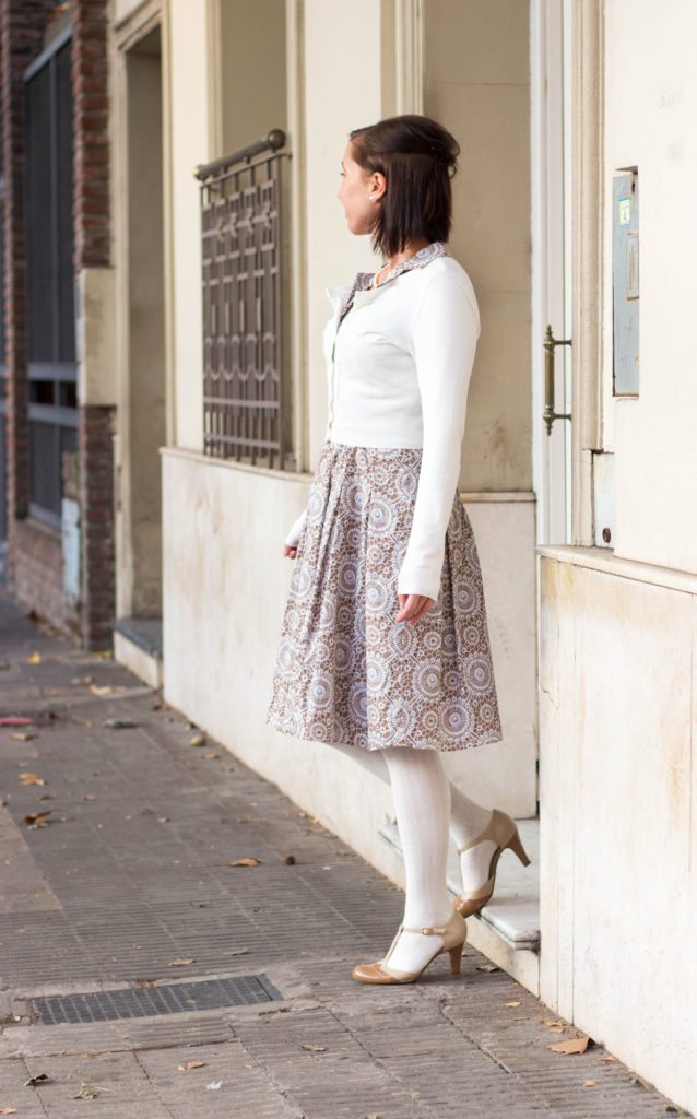 A woman poses on a city street wearing a patterned tan and white dress under a long-sleeve white cardigan, white knit tights, and nude heels.