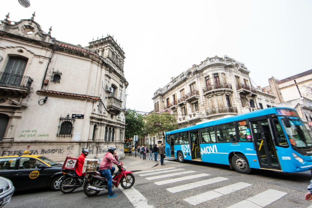 View of an Argentinian street with a bright blue bus and two motorcyclists passing by. 